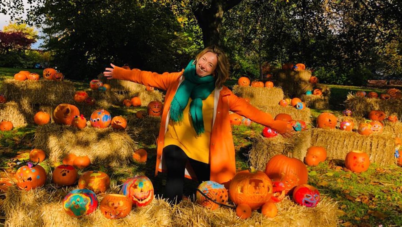 A person with a pumpkin for a head stands in Glasgow Botanic Gardens on an autumn day. They are surrounded by small decorated pumpkins.